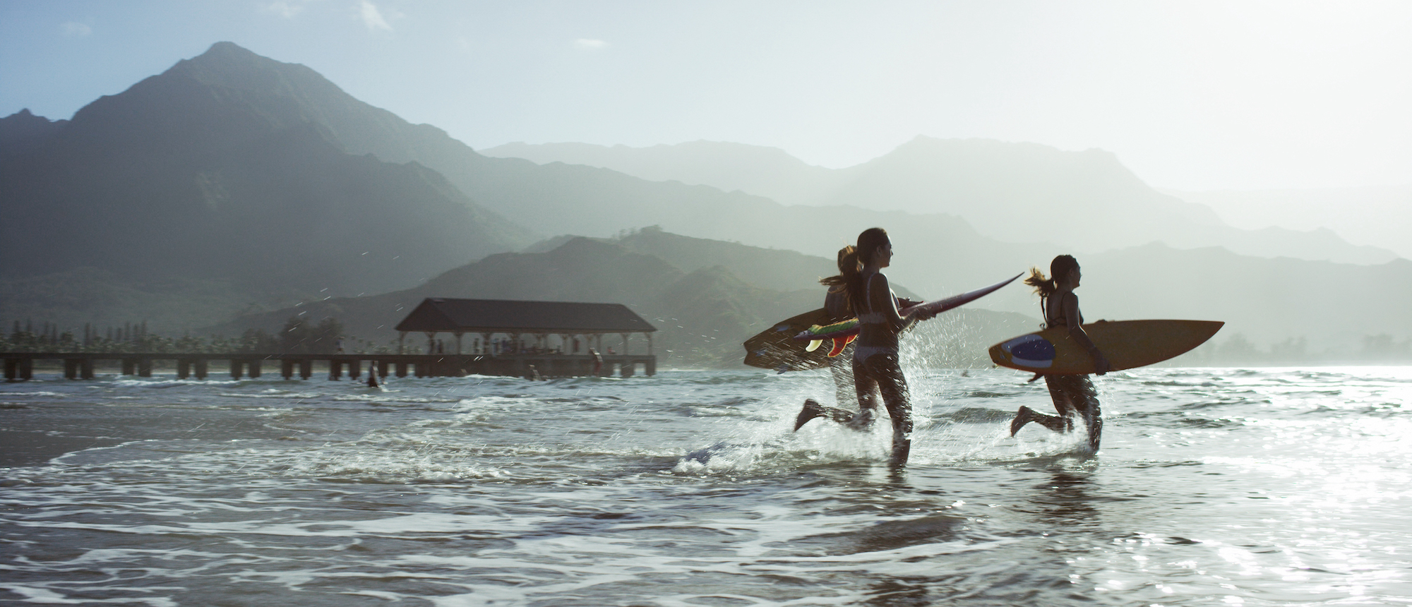 Three people on a beach, running into the ocean with surboards in a tropical climate, with mountains in the background.